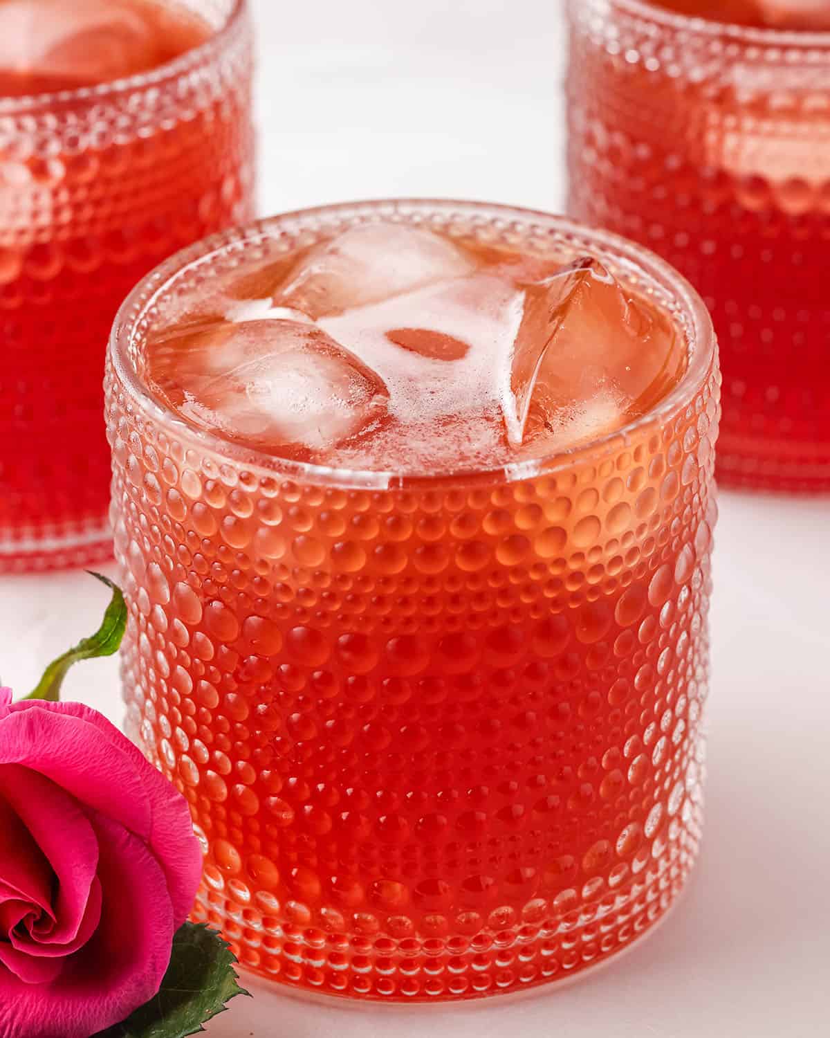 A vintage looking glass with red rose kombucha and ice in it, on a white counter, with a deep pink rose laying aside it. with other glasses of rose kombucha in the background. 